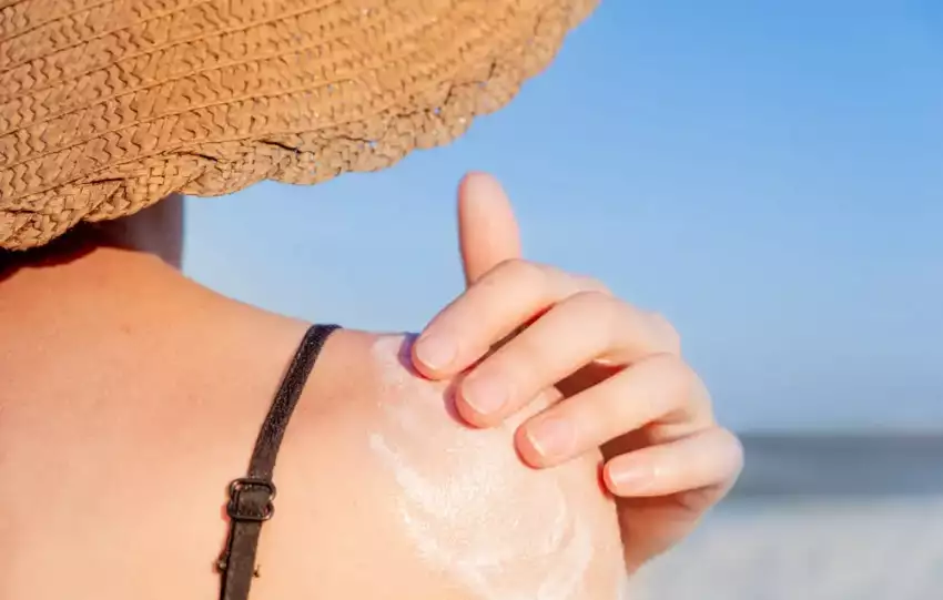 mulher passando protetor solar no ombro e usando chapéu para se prevenir dos perigos da exposição em excesso ao sol