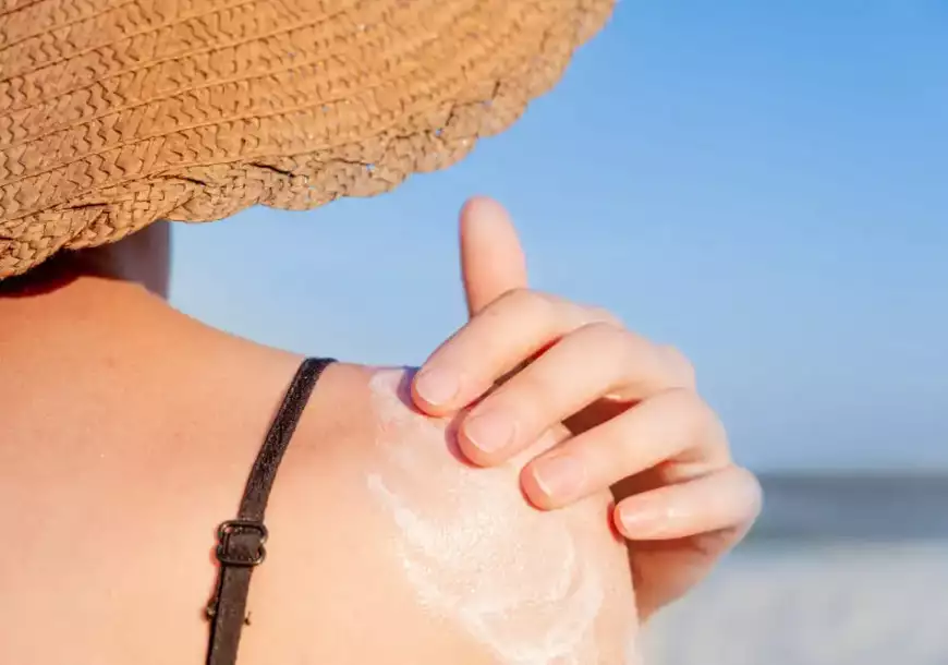mulher passando protetor solar no ombro e usando chapéu para se prevenir dos perigos da exposição em excesso ao sol