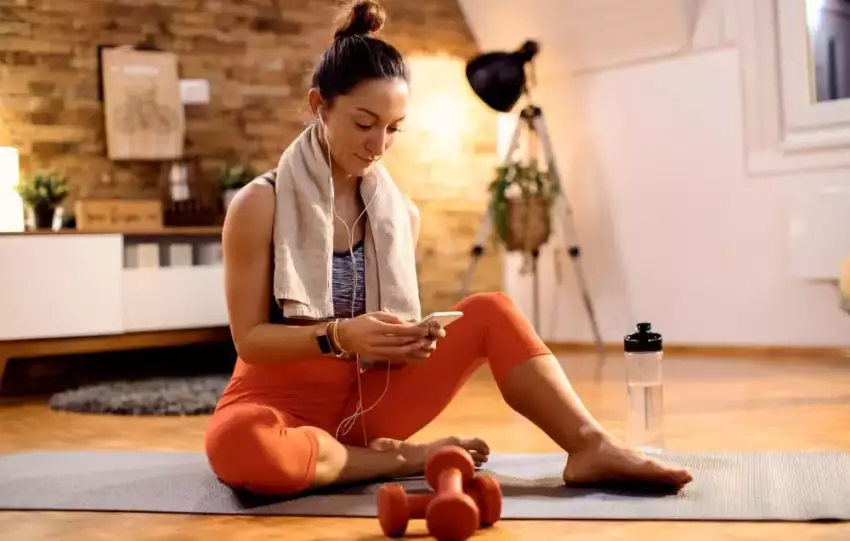 mulher-branca-usando-legging-laranja-com-toalha-em-volta-do-pescoco-escolhendo-uma-musica-para-tocar-em-seu-fone-para-fazer-exercicio-em-casa